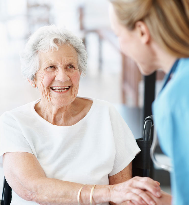 respite-care Elderly woman in a white shirt smiling warmly at a caregiver in blue scrubs during a visit