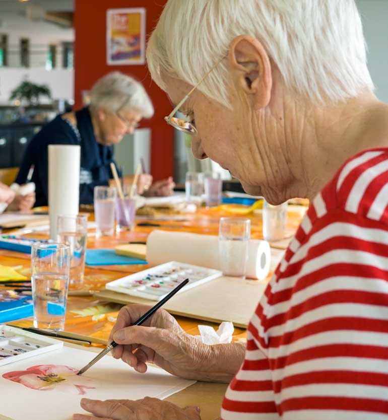 senior-painting Senior woman painting watercolor at a table with other seniors in an art setting.