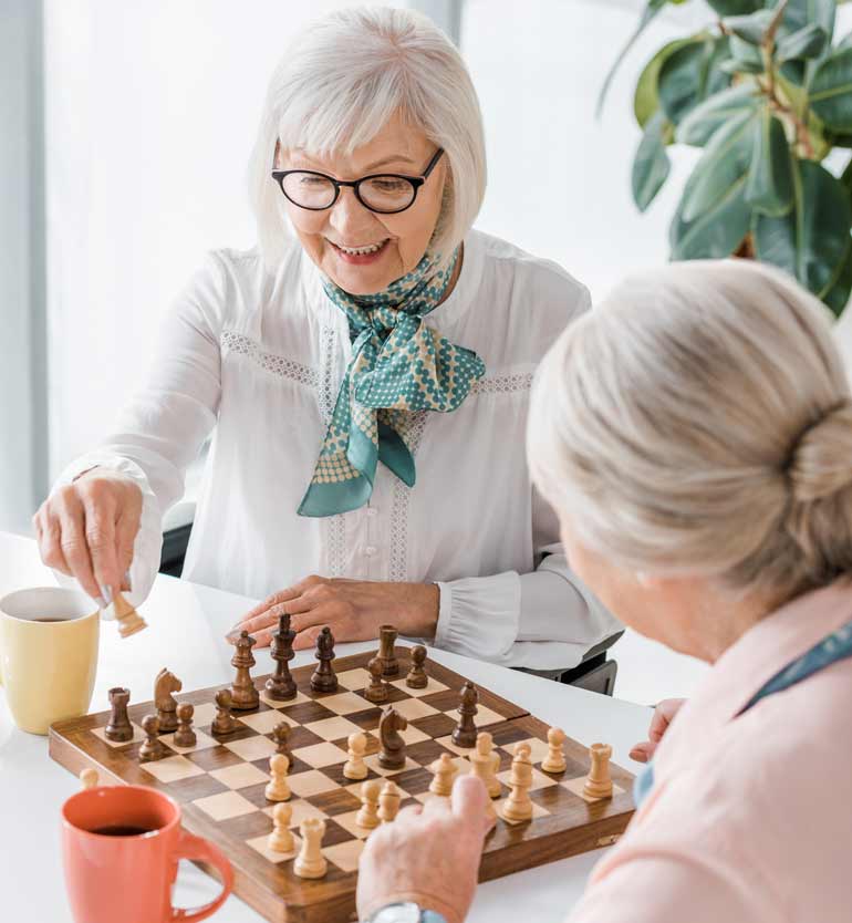 seniors-playing-chess Two senior women happily engaged in a chess game at a bright indoor setting with coffee mugs nearby.