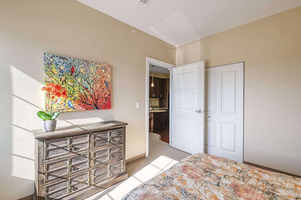 Sunlit bedroom with dresser, bright painting, and vase of orange flowers.